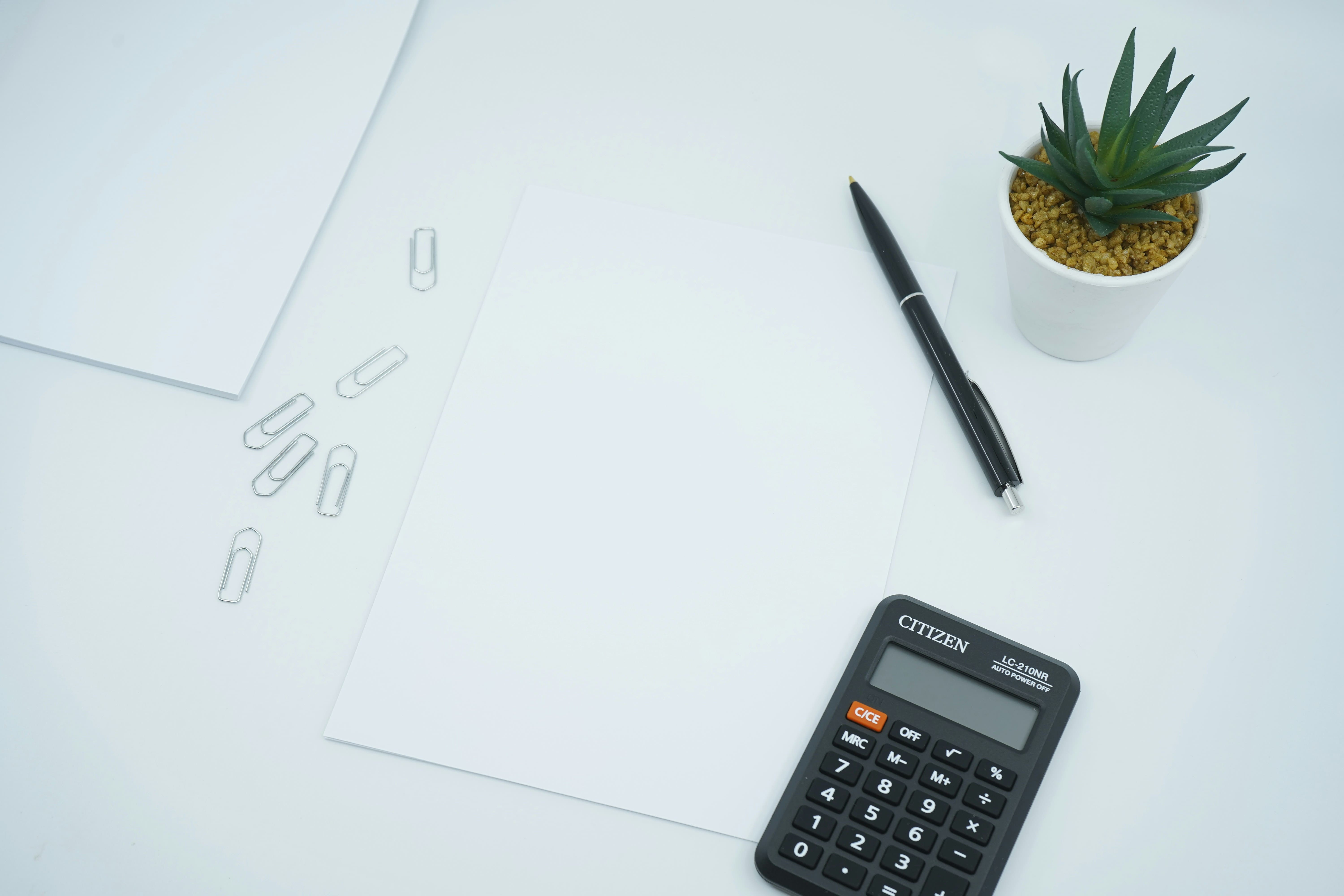 A desk with a calculator, pen, paper clips and a small plant